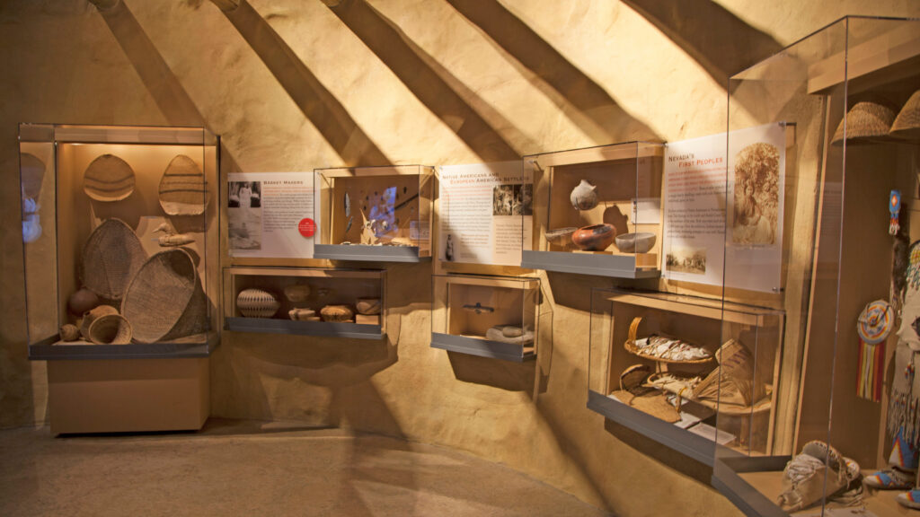 A museum exhibit displays Native American baskets, pottery, and artifacts in glass cases against tan walls, with descriptive signs and soft lighting casting shadows across the room.