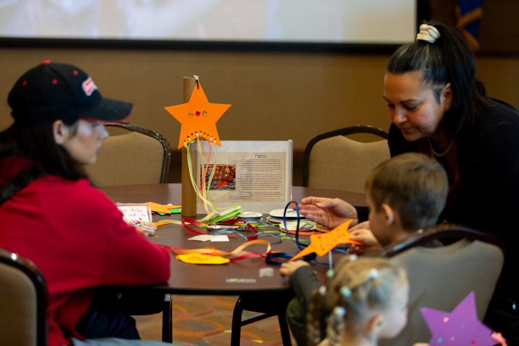Two adults and two children sit at a table doing a star-themed craft with colored paper and ribbons. The table is covered with supplies and finished stars. A sign with instructions is visible in the background.