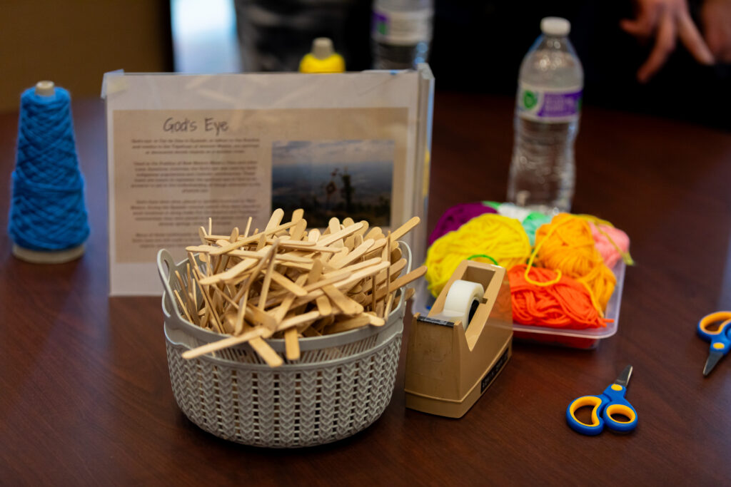 Craft supplies on a table, including popsicle sticks in a basket, colorful yarn, a tape dispenser, scissors, water bottles, and a printed instruction sheet titled Gods Eye..