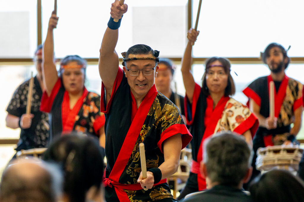 A group of people in traditional attire energetically play taiko drums in front of an audience, with one drummer raising a stick in the air.