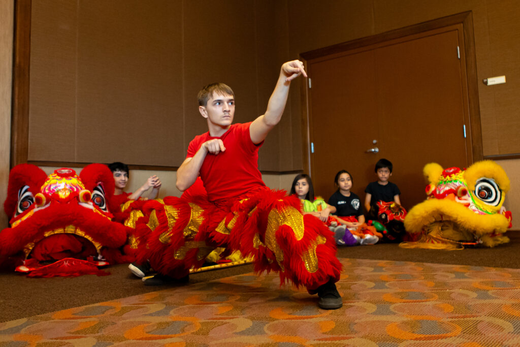 A person in a red and gold lion dance costume poses in a martial arts stance, while children sit in the background with lion dance heads on the floor beside them.