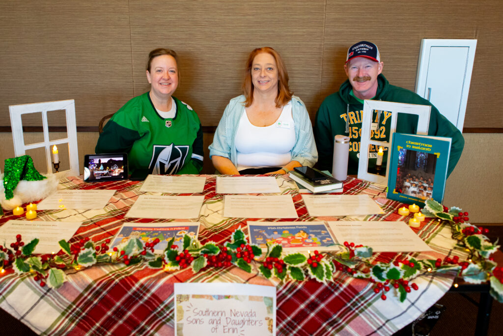 Three people sit behind a festive table with holiday decorations, papers, framed photos, and a Southern Nevada Sons and Daughters of Erin sign, smiling at the camera in a warmly lit indoor setting.